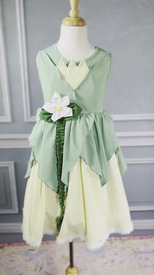 A young girl poses in the Frog Princess Twirl Dress . She wears a green and yellow dress with a magnolia accent at the waist. The light green top and leaf shaped skirt are overlayed on a light yellow skirt with white eyelet trim at the bottom.  The dress is sleeveless and a comfortable cotton. 