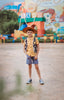 A young boy stands dressed in a sheriff cowboy outfit from the Toyland Collection. A yellow and red checkered short sleeve shirt featuring black and white cow print vest. A yellow sheriff badge is sewn on the right chest of the vest. The collar shirt features buttons in the middle and one on each sleeve.