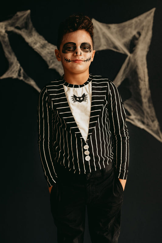 A child wears black and white face paint resembling a skeleton, with dark eye circles and a stitched mouth. Dressed in a comfortable poly-knit fabric jacket over "The Pumpkin King Shirt," which features a spooky t-shirt adorned with a small bat motif, the child stands against a dark background with spider webs.