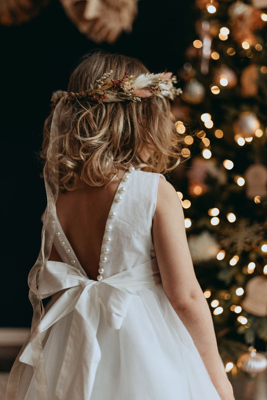 A young girl in a Pearl Accent gown in white, featuring a bow at the back. Woven cotton bodice and large pearl beads line the neck and back edge! A full tulle skirt with tea length hemline and modest open back.