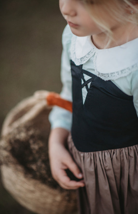 A young child wearing The Fairy Godmother Collection Sleeping Beauty dress with a white collar and lace trim. The black bodice and light brown skirt are woven cotton.  The dress structure is fitted in the top and flares out as the waist. 