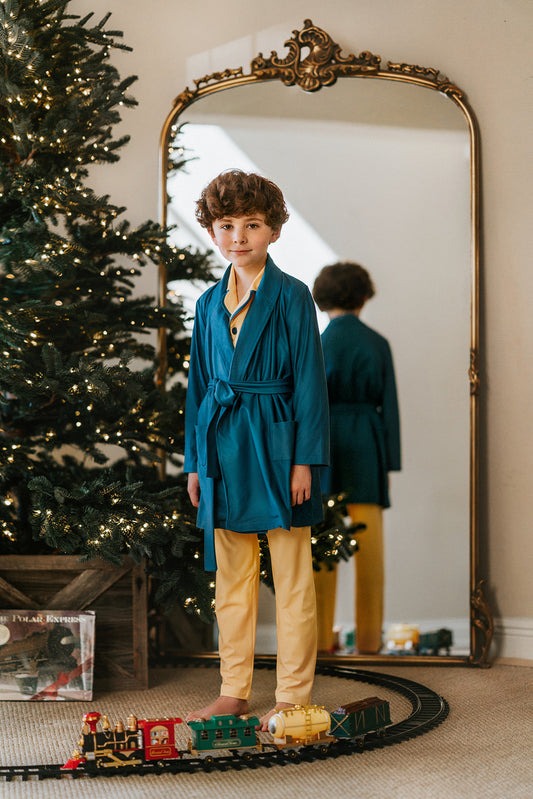 A young boy is dressed in a bright yellow classic 2 piece button up pajama set.  The top is has blue green piping and buttons.   He has a long sleeve blue green robe tied at the waist. 