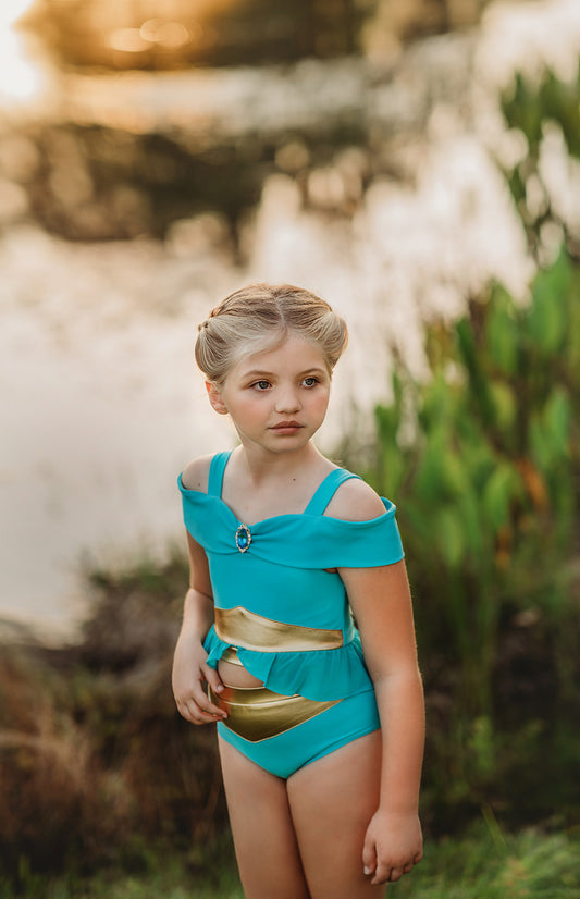 A young girl is wearing a bight teal and gold 2pc swim set.  The top has open shoulders and straps.  At the hem is a gold band and teal ruffles. There is a teal blue and gold jewel at the neckline.  The bottom is a modest cut with gold waist and teal. 