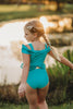 A young girl with blonde hair in a long braid stands facing a pond, wearing an Arabian Princess Two Piece Swimsuit in turquoise adorned with ruffles. The setting sun reflects on the water, creating a warm, golden glow in the background. She is surrounded by lush greenery.