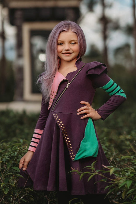 A young girl with long, purple hair, often referred to as the Mischievous Miss, smiles while standing outdoors. She is wearing a Mischievous Miss dress made of jersey fabric with colorful accents, including green and pink patches and gold studs. The background features greenery and a blurred structure.