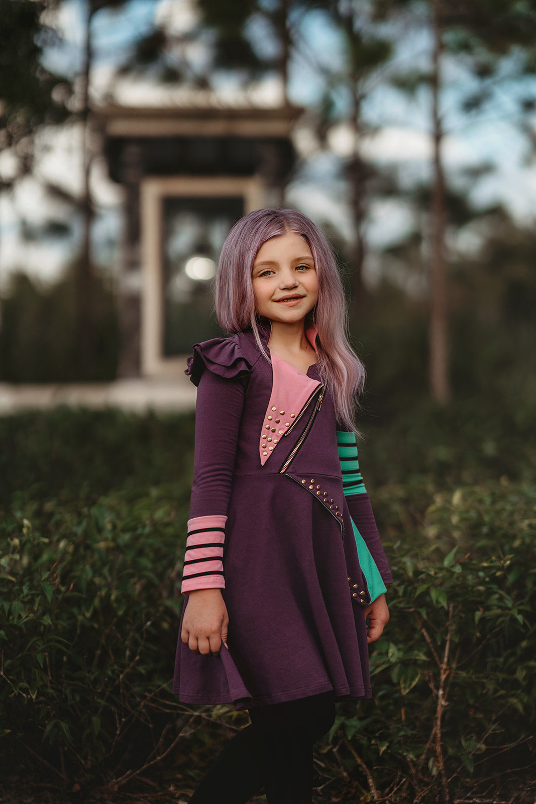 A young girl  is wearing a Mischievous Miss dress made of jersey fabric with colorful accents, including green and pink patches and gold studs. There is a asymmetrical zipper and ruffle on the shoulder the the long sleeves.  