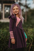 A young girl with long, purple hair, often referred to as the Mischievous Miss, smiles while standing outdoors. She is wearing a Mischievous Miss dress made of jersey fabric with colorful accents, including green and pink patches and gold studs. The background features greenery and a blurred structure.