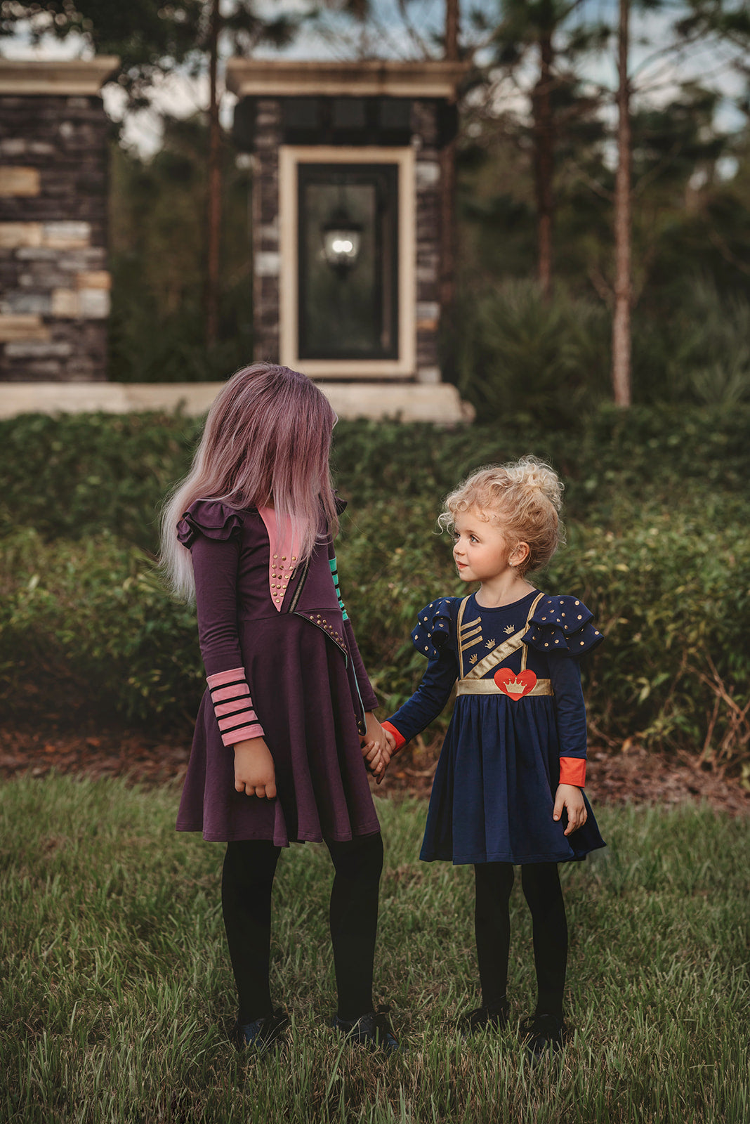 A young girl with long, purple hair, often referred to as the Mischievous Miss, smiles while standing outdoors. She is wearing a Mischievous Miss dress made of jersey fabric with colorful accents, including green and pink patches and gold studs. The background features greenery and a blurred structure.