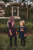 A young girl with long, purple hair, often referred to as the Mischievous Miss, smiles while standing outdoors. She is wearing a Mischievous Miss dress made of jersey fabric with colorful accents, including green and pink patches and gold studs. The background features greenery and a blurred structure.