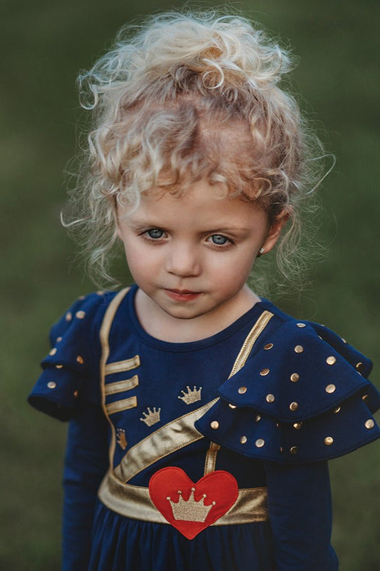 A young girl is wearing a long sleeve navy blue twirl dress. The sleeves have two large flutter ruffles at the shoulder with metal gold dots.  The chest is adorned with gold detail and crowns.  A red heart with a gold crown in the center is on the lower left chest. 