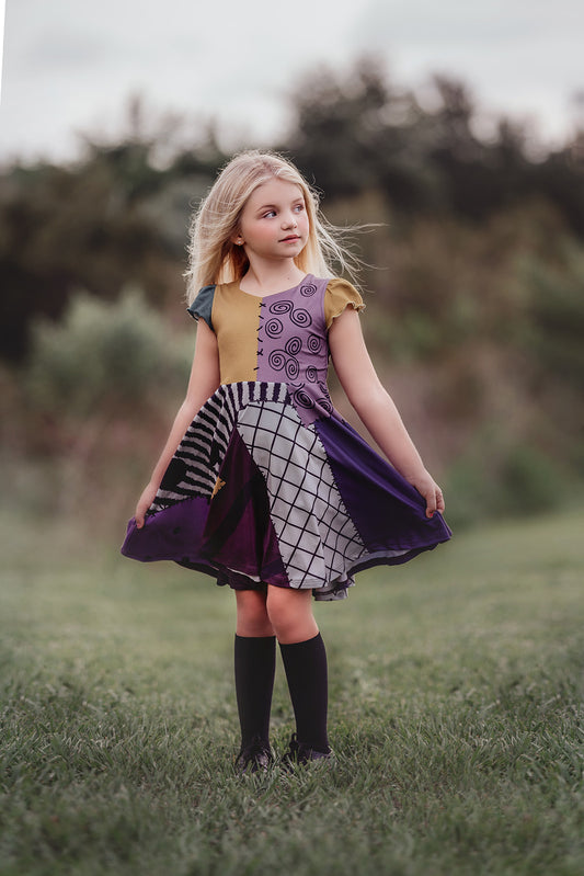 A young girl is wearing "The Pumpkin King's Friend" dress, its colorful patchwork in purple, black, and mustard yellow made of soft poly-spandex blend fabric. Perfect for spooky season!