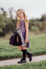 A young girl with long blonde hair is standing on a grassy field, wearing "The Pumpkin King's Friend" dress, its colorful patchwork in purple, black, and green made of soft poly-spandex blend fabric. She pairs the dress with black knee-high socks and looks off to the side while holding the edges of her dress. Trees stand in the background.