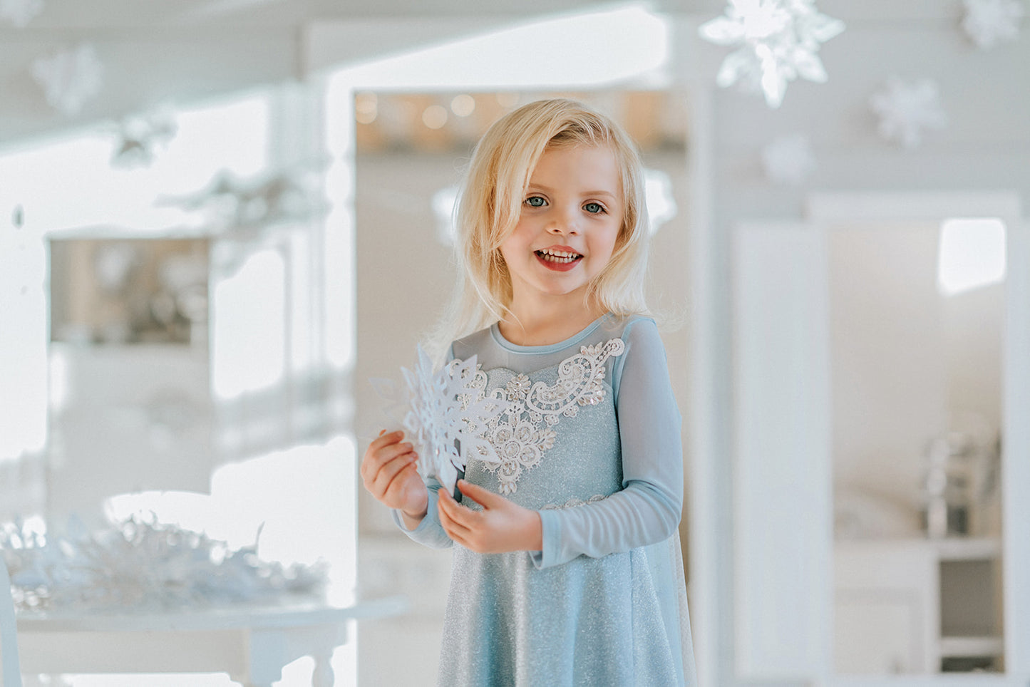 A young girl is dressed in the sparkly light blue Portrait Collection Ice Queen Gown adorned with intricate custom  lace at the neckline with rhinestones. A light blue cape with silver sparkle snowflakes is buttoned to the back of the gown. 