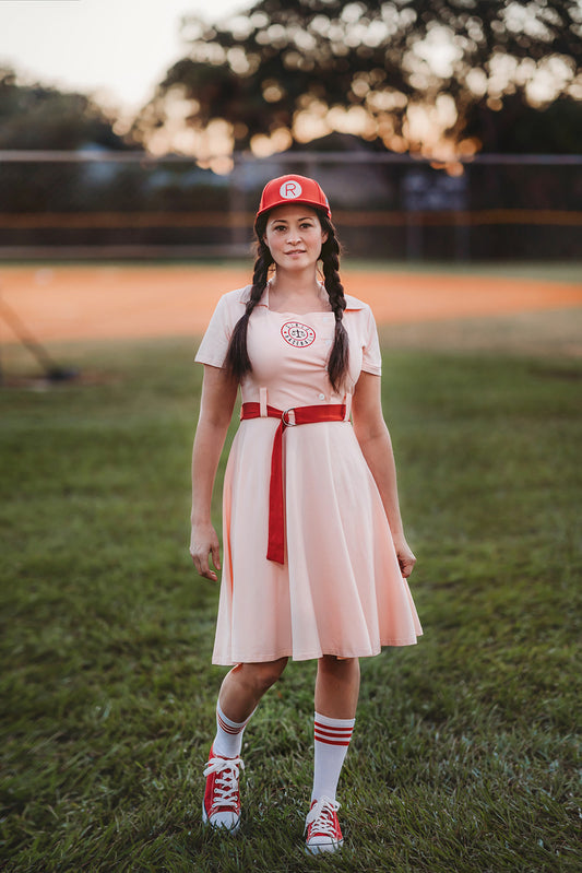 A person wearing the Adult Vintage Baseball Dress, styled with a red belt, a red cap featuring an "R," striped socks, and red sneakers, stands on grass. The outfit is crafted from soft cotton jersey knit fabric and is available in adult sizes. A blurred baseball field and trees form the background.