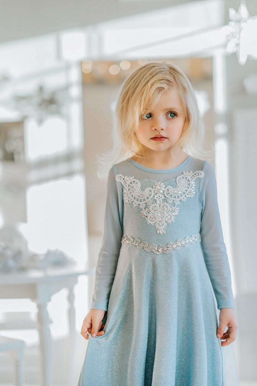 A young girl is dressed in the sparkly light blue Portrait Collection Ice Queen Gown adorned with intricate custom  lace at the neckline with rhinestones. A light blue cape with silver sparkle snowflakes is buttoned to the back of the gown. 