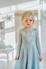 A young girl is dressed in the sparkly light blue Portrait Collection Ice Queen Gown adorned with intricate custom  lace at the neckline with rhinestones. A light blue cape with silver sparkle snowflakes is buttoned to the back of the gown. 