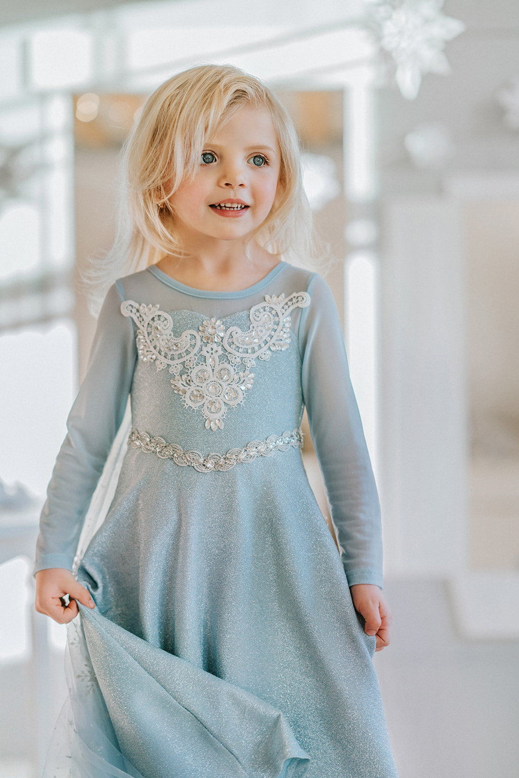 A young girl is dressed in the sparkly light blue Portrait Collection Ice Queen Gown adorned with intricate custom  lace at the neckline with rhinestones. A light blue cape with silver sparkle snowflakes is buttoned to the back of the gown. 