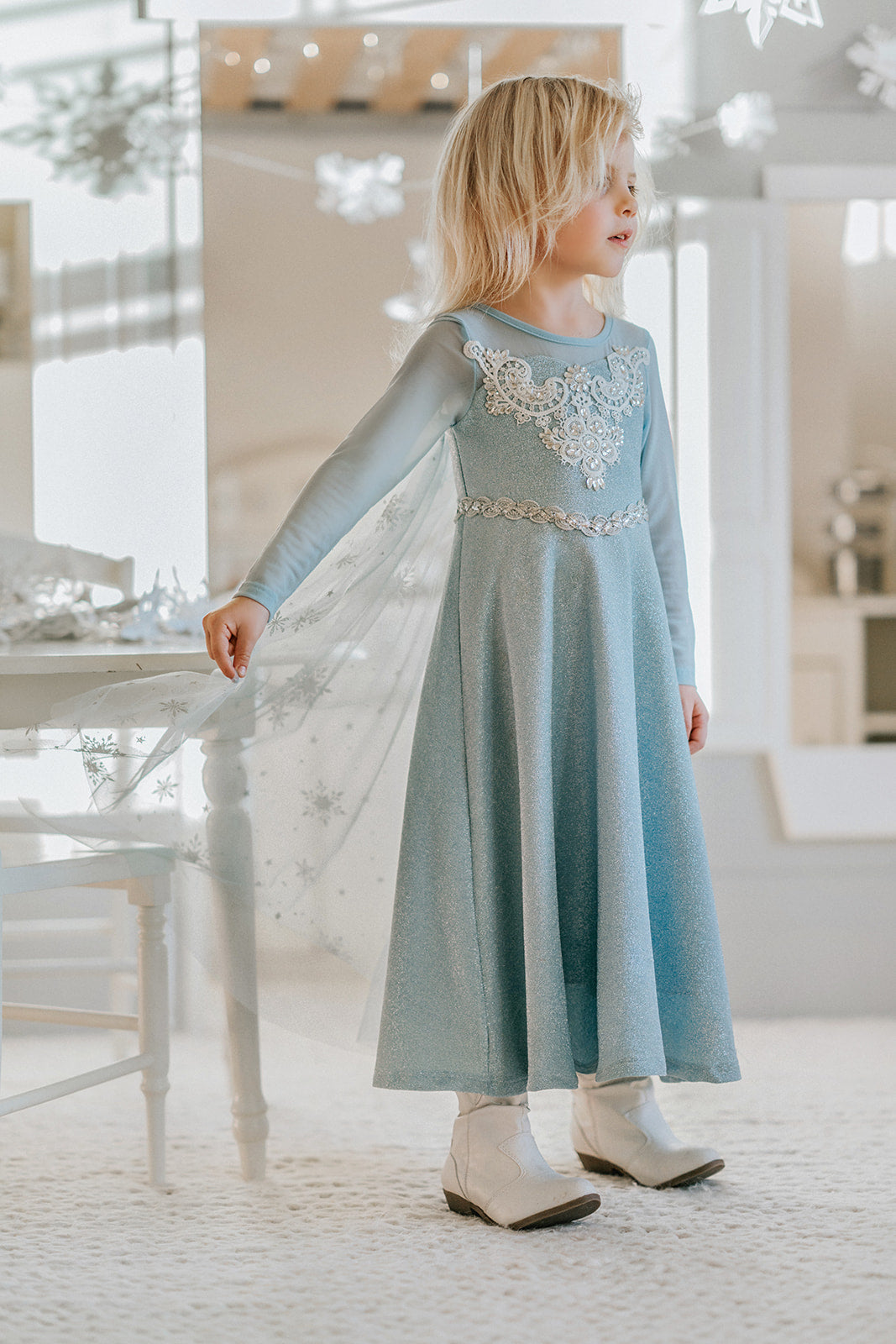 A young girl is dressed in the sparkly light blue Portrait Collection Ice Queen Gown adorned with intricate custom  lace at the neckline with rhinestones. A light blue cape with silver sparkle snowflakes is buttoned to the back of the gown. 