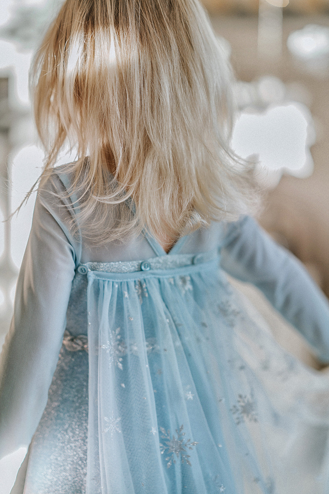 A young girl is dressed in the sparkly light blue Portrait Collection Ice Queen Gown adorned with intricate custom  lace at the neckline with rhinestones. A light blue cape with silver sparkle snowflakes is buttoned to the back of the gown. 