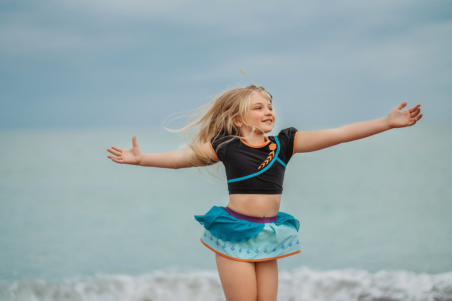 A young girl is wearing a 2 piece long sleeve rash guard swim set. The top is black with turquoise and goldenrod accents. The bottom has a turquoise ruffle over a mint teal skirt with goldenrod hem accent. 