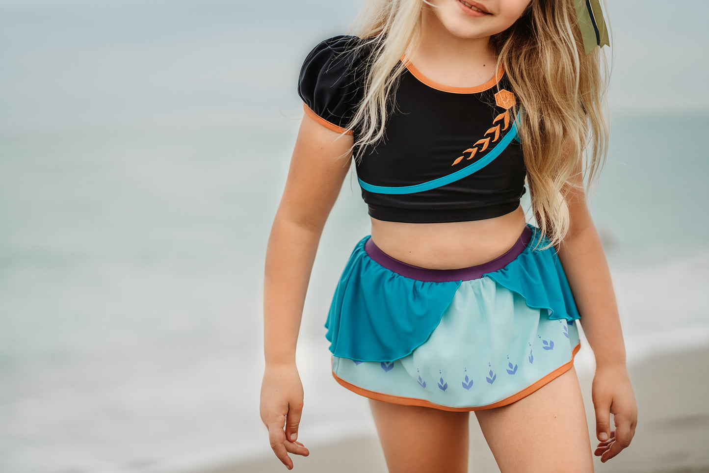 A young girl is wearing a 2 piece long sleeve rash guard swim set. The top is black with turquoise and goldenrod accents. The bottom has a turquoise ruffle over a mint teal skirt with goldenrod hem accent. 