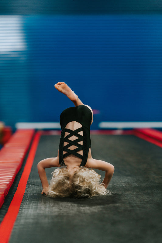 A young girl is wearing the Mae Leotard in black.  The classic cut leo has a criss-cross straps over the back cut out and eyelet ruffles along the edge. The leo is SPF 50 and could be worn as swimwear also. 