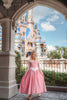 A young girl in an exquisite Portrait Collection Sleeping Beauty gown, complete with a sparkling jeweled crown, stands in front of a stone castle backdrop. Smiling, she holds out the sides of her elegant dress. The whimsical lighting enhances the fairytale ambiance, creating an enchanting scene perfect for the Portrait Collection.