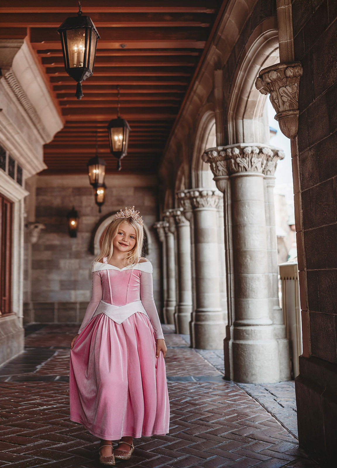 A young girl in an exquisite Portrait Collection Sleeping Beauty gown, complete with a sparkling jeweled crown, stands in front of a stone castle backdrop. Smiling, she holds out the sides of her elegant dress. The whimsical lighting enhances the fairytale ambiance, creating an enchanting scene perfect for the Portrait Collection.