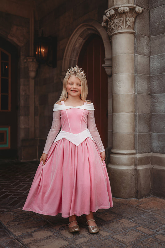 A young girl in an exquisite Portrait Collection Sleeping Beauty gown poses in a castle setting.  The pink stretch velvet gown has an off the shoulder neckline with two straps.  The long sleeves are light pink mesh.  The dress has sewn in tulle and a cotton liner to add fullness in the skirt. 