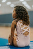 A young girl is wearing the Mae Leotard in light pink.  The classic cut leo has a criss-cross straps over the back cut out and eyelet ruffles along the edge. The leo is SPF 50 and could be worn as swimwear also. 