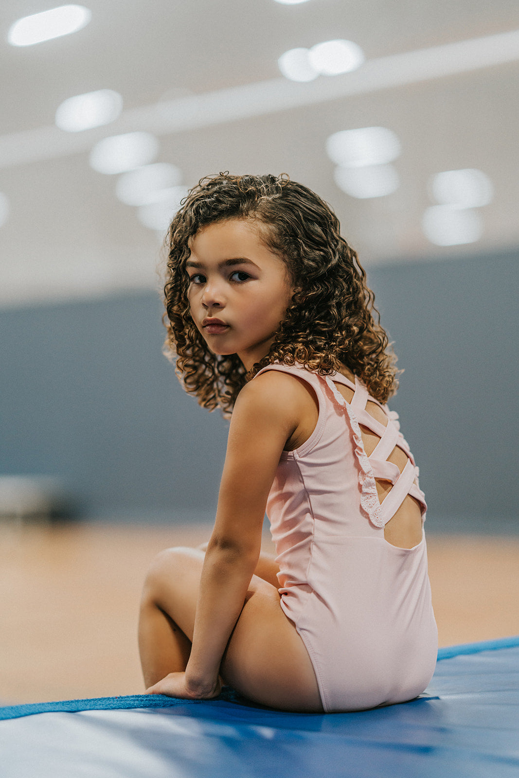 A young girl is wearing the Mae Leotard in light pink.  The classic cut leo has a criss-cross straps over the back cut out and eyelet ruffles along the edge. The leo is SPF 50 and could be worn as swimwear also. 