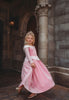 A young girl in an exquisite Portrait Collection Sleeping Beauty gown, complete with a sparkling jeweled crown, stands in front of a stone castle backdrop. Smiling, she holds out the sides of her elegant dress. The whimsical lighting enhances the fairytale ambiance, creating an enchanting scene perfect for the Portrait Collection.