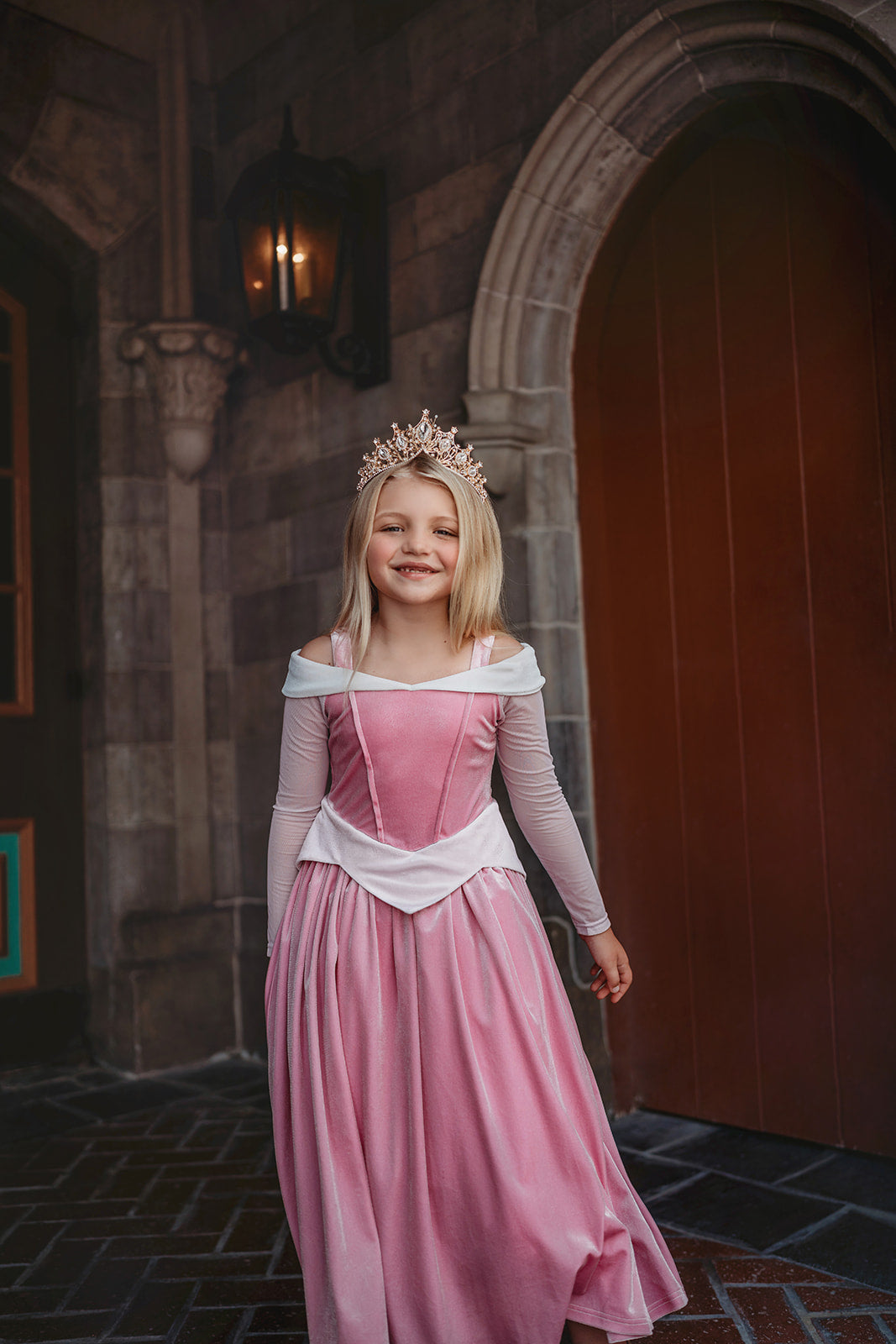 A young girl in an exquisite Portrait Collection Sleeping Beauty gown, complete with a sparkling jeweled crown, stands in front of a stone castle backdrop. Smiling, she holds out the sides of her elegant dress. The whimsical lighting enhances the fairytale ambiance, creating an enchanting scene perfect for the Portrait Collection.