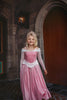 A young girl in an exquisite Portrait Collection Sleeping Beauty gown, complete with a sparkling jeweled crown, stands in front of a stone castle backdrop. Smiling, she holds out the sides of her elegant dress. The whimsical lighting enhances the fairytale ambiance, creating an enchanting scene perfect for the Portrait Collection.