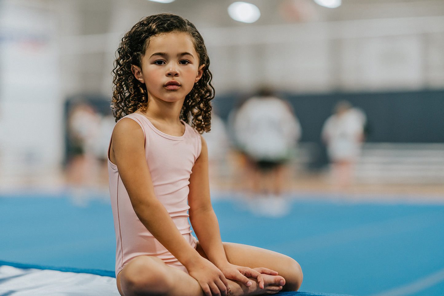 A young girl is wearing the Mae Leotard in light pink.  The classic cut leo has a criss-cross straps over the back cut out and eyelet ruffles along the edge. The leo is SPF 50 and could be worn as swimwear also. 