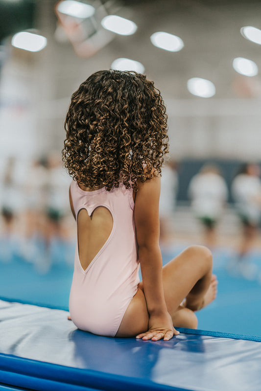 A young girl with curly hair sits on a gymnastics mat, facing away from the camera, in a gymnasium. She is wearing the Love Bug Leotard, a pink gymnastics outfit with an elegant heart cutout back. The background is slightly blurred, showing other people and gym equipment.