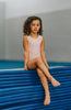 A young girl is wearing the Mae Leotard in light pink.  The classic cut leo has a criss-cross straps over the back cut out and eyelet ruffles along the edge. The leo is SPF 50 and could be worn as swimwear also. 