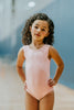 A young girl is wearing the Love Bug Leotard, a pink gymnastics outfit with an elegant heart cutout back. The background is slightly blurred, showing other people and gym equipment.