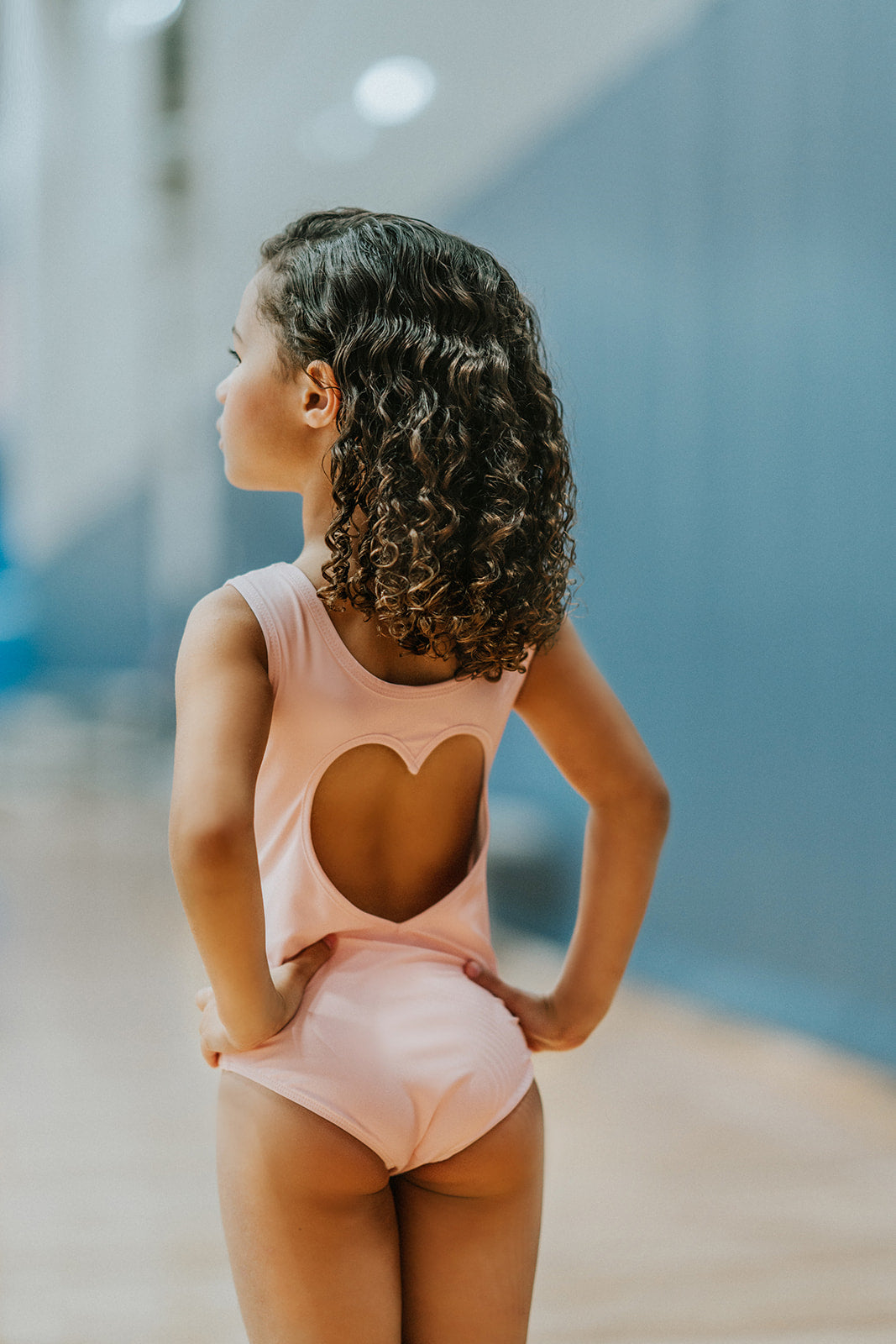 A young girl is wearing the Love Bug Leotard, a pink gymnastics outfit with an elegant heart cutout back. The background is slightly blurred, showing other people and gym equipment.