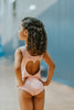 A young girl is wearing the Love Bug Leotard, a pink gymnastics outfit with an elegant heart cutout back. The background is slightly blurred, showing other people and gym equipment.