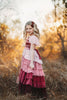 A young girl is wearing the Yule Love all of the ruffles Maxi Dress in shades of pink and red. The soft pink flutter sleeves flow over her shoulder.  The bodice is a pink mauve and leads into cascading layers chiffon that start with dusty pink and flow down into a deep maroon. 