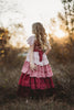 A young girl is wearing the Yule Love all of the ruffles Maxi Dress in shades of pink and red. The soft pink flutter sleeves flow over her shoulder.  The bodice is a pink mauve and leads into cascading layers chiffon that start with dusty pink and flow down into a deep maroon. 