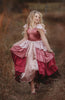 A young girl is wearing the Yule Love all of the ruffles Maxi Dress in shades of pink and red. The soft pink flutter sleeves flow over her shoulder.  The bodice is a pink mauve and leads into cascading layers chiffon that start with dusty pink and flow down into a deep maroon. 