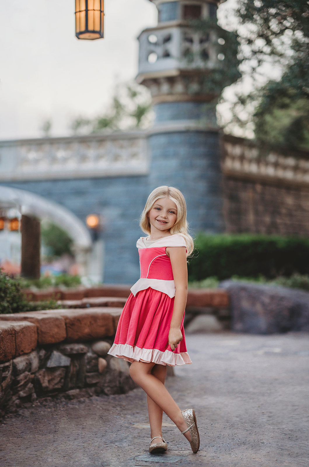 A young girl is wearing an all cotton bright pink princess twirl dress. The neckline has nude mesh and light pink sits off the shoulder.  The bright pink bodice has light pink piping and leads into a light pink handkerchief band.  The skirt is flowy and bright pink.  A light pink ruffle at the hem line adds a beautiful twirl. 