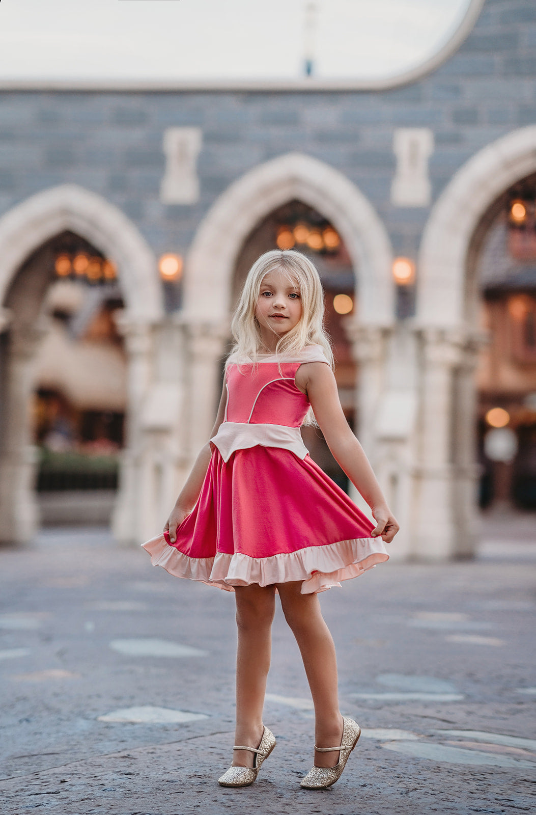 A young girl is wearing an all cotton bright pink princess twirl dress. The neckline has nude mesh and light pink sits off the shoulder.  The bright pink bodice has light pink piping and leads into a light pink handkerchief band.  The skirt is flowy and bright pink.  A light pink ruffle at the hem line adds a beautiful twirl. 