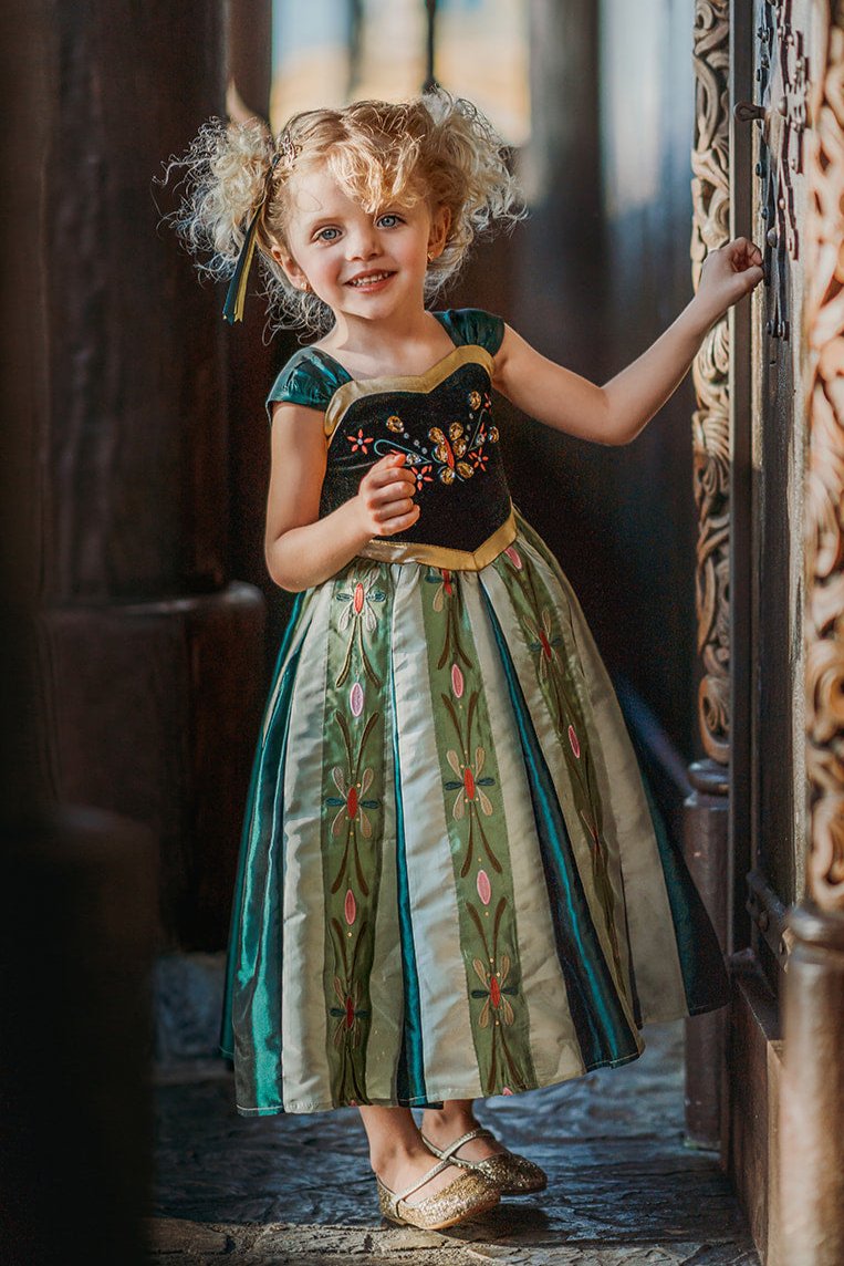 A young girl is wearing a Portrait Collection Coronation Ice Princess Gown inspired by Princess Anna from the movie "Frozen." The dress showcases a black velvet bodice with custom embroidery and a skirt adorned with green and gold accents. The skirt is shimmering in shades of green with embroidered designs along the panels.  There is tulle with cotton lining underneath to add fullness to the skirt. 