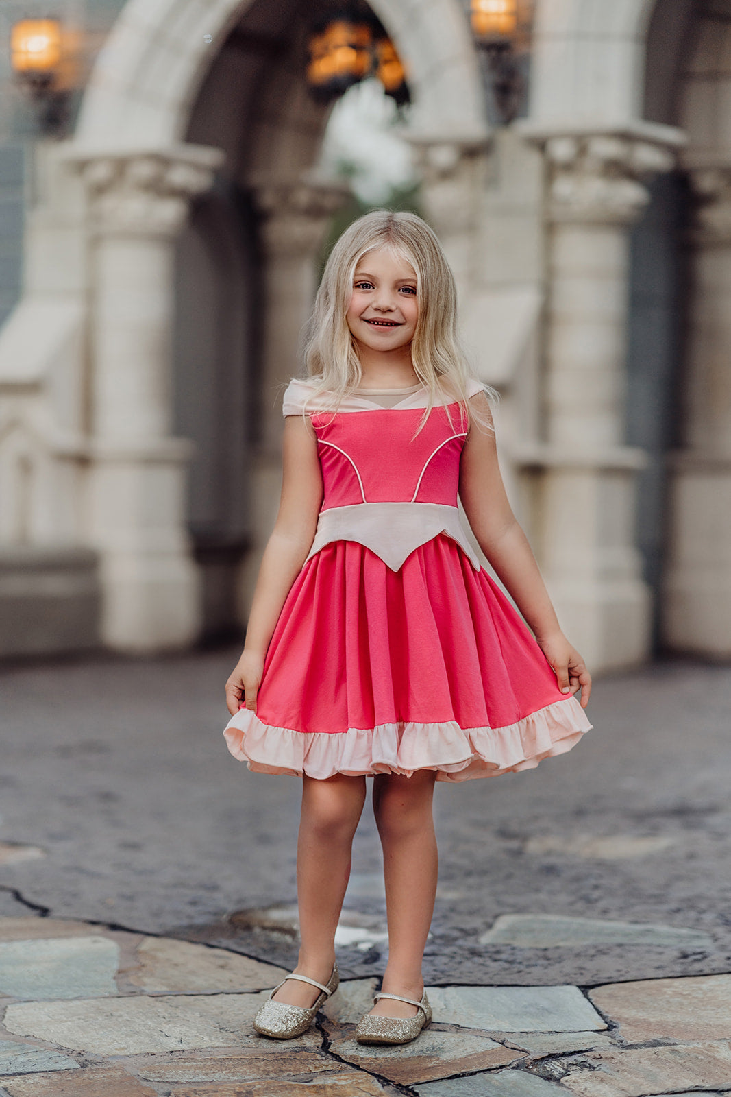 A young girl is wearing an all cotton bright pink princess twirl dress. The neckline has nude mesh and light pink sits off the shoulder.  The bright pink bodice has light pink piping and leads into a light pink handkerchief band.  The skirt is flowy and bright pink.  A light pink ruffle at the hem line adds a beautiful twirl. 