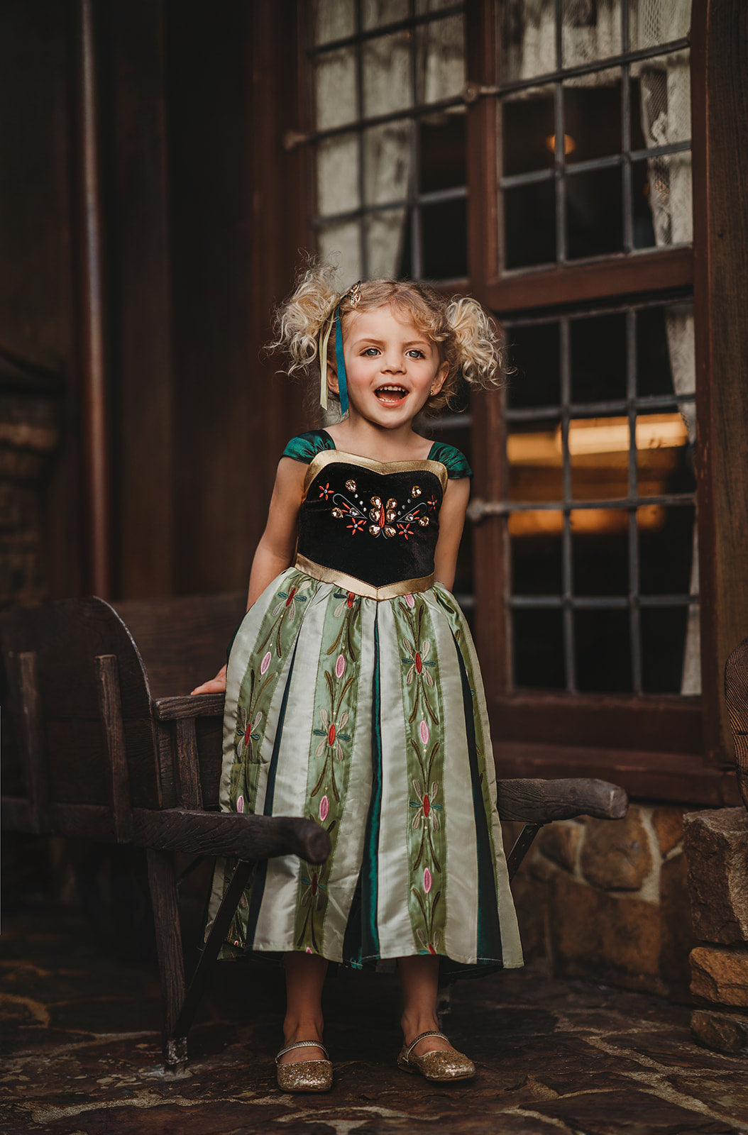 A young girl is wearing a Portrait Collection Coronation Ice Princess Gown inspired by Princess Anna from the movie "Frozen." The dress showcases a black velvet bodice with custom embroidery and a skirt adorned with green and gold accents. The skirt is shimmering in shades of green with embroidered designs along the panels.  There is tulle with cotton lining underneath to add fullness to the skirt. 