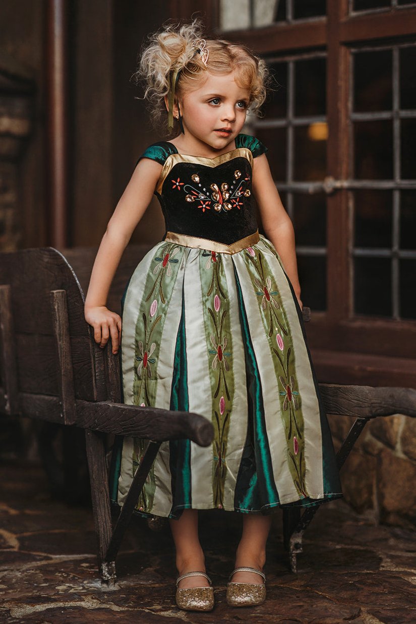 A young girl is wearing a Portrait Collection Coronation Ice Princess Gown inspired by Princess Anna from the movie "Frozen." The dress showcases a black velvet bodice with custom embroidery and a skirt adorned with green and gold accents. The skirt is shimmering in shades of green with embroidered designs along the panels.  There is tulle with cotton lining underneath to add fullness to the skirt. 