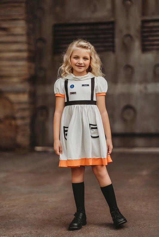 A young girl is wearing a white cotton twirl dress with a hood to resemble an astronaut space suit. The puff sleeves and skirt are trimmed in orange.  There is a faux strap over each shoulder.  The dress has two pockets on the skirt and on the bodice three space inspired patches. 